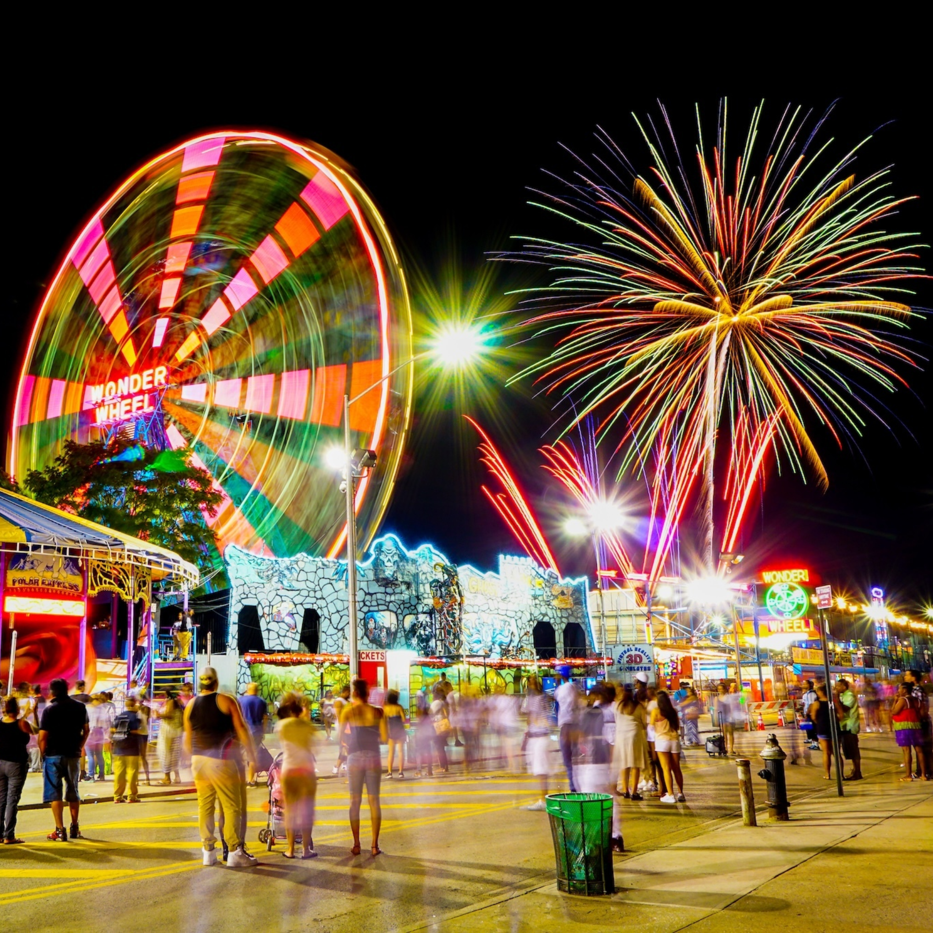 NYC Wonder Wheel amusement park at night with colorful lights, fireworks, and crowds under neon signs, festive carnival atmosphere featured in daily things to do in NYC any day