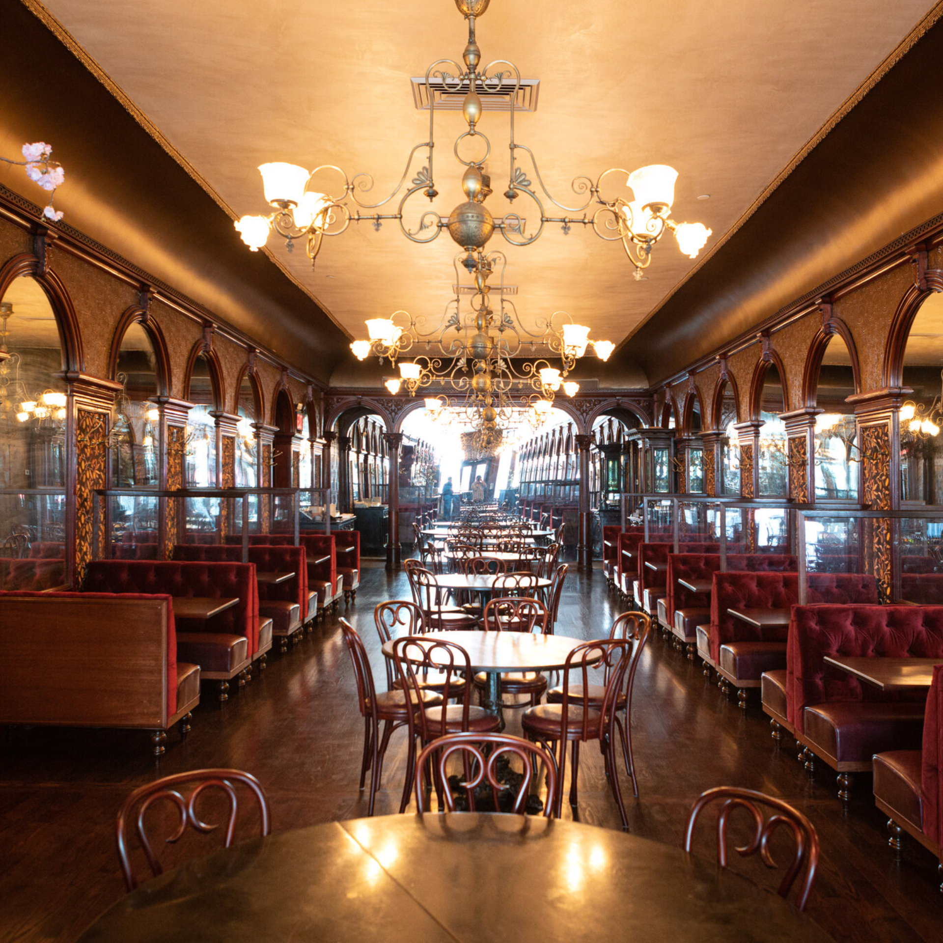 Vintage restaurant interior with ornate chandeliers, red velvet booths, arched mirrors, carved wood walls, round tables, curved‑back chairs, polished dark floors