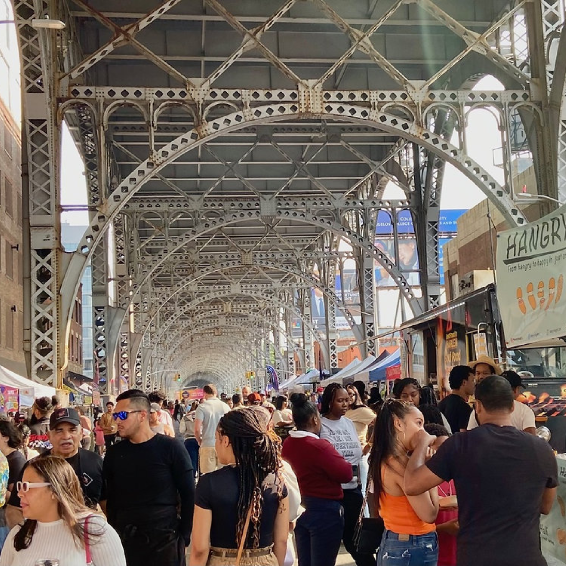 NYC street market under steel bridge with vendor stalls, food trucks, and crowds browsing, lively urban community event featured in daily things to do in NYC any day