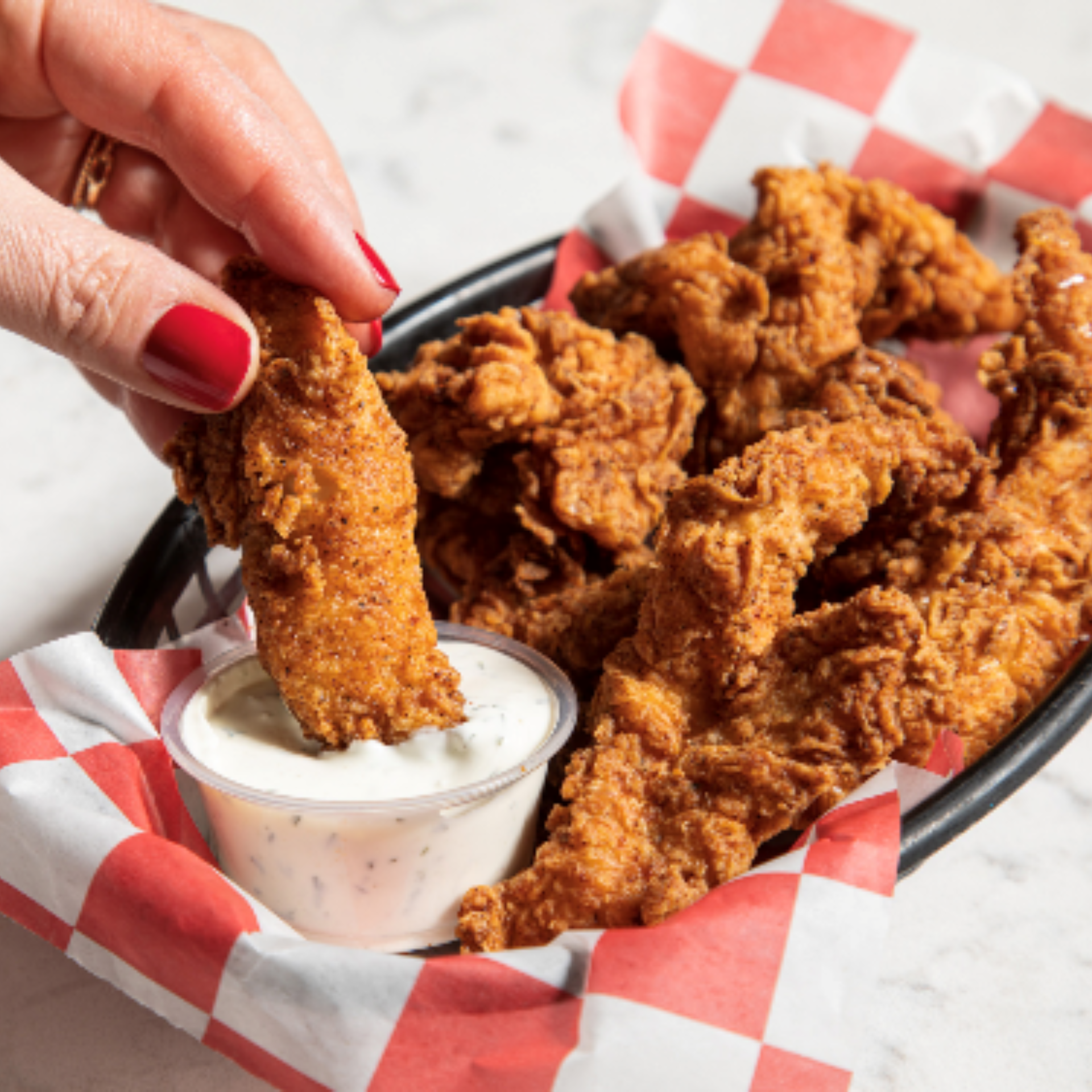 Basket of crispy fried chicken tenders on checkered paper with hand dipping into creamy sauce, NYC comfort food favorite featured in daily things to do in NYC any day