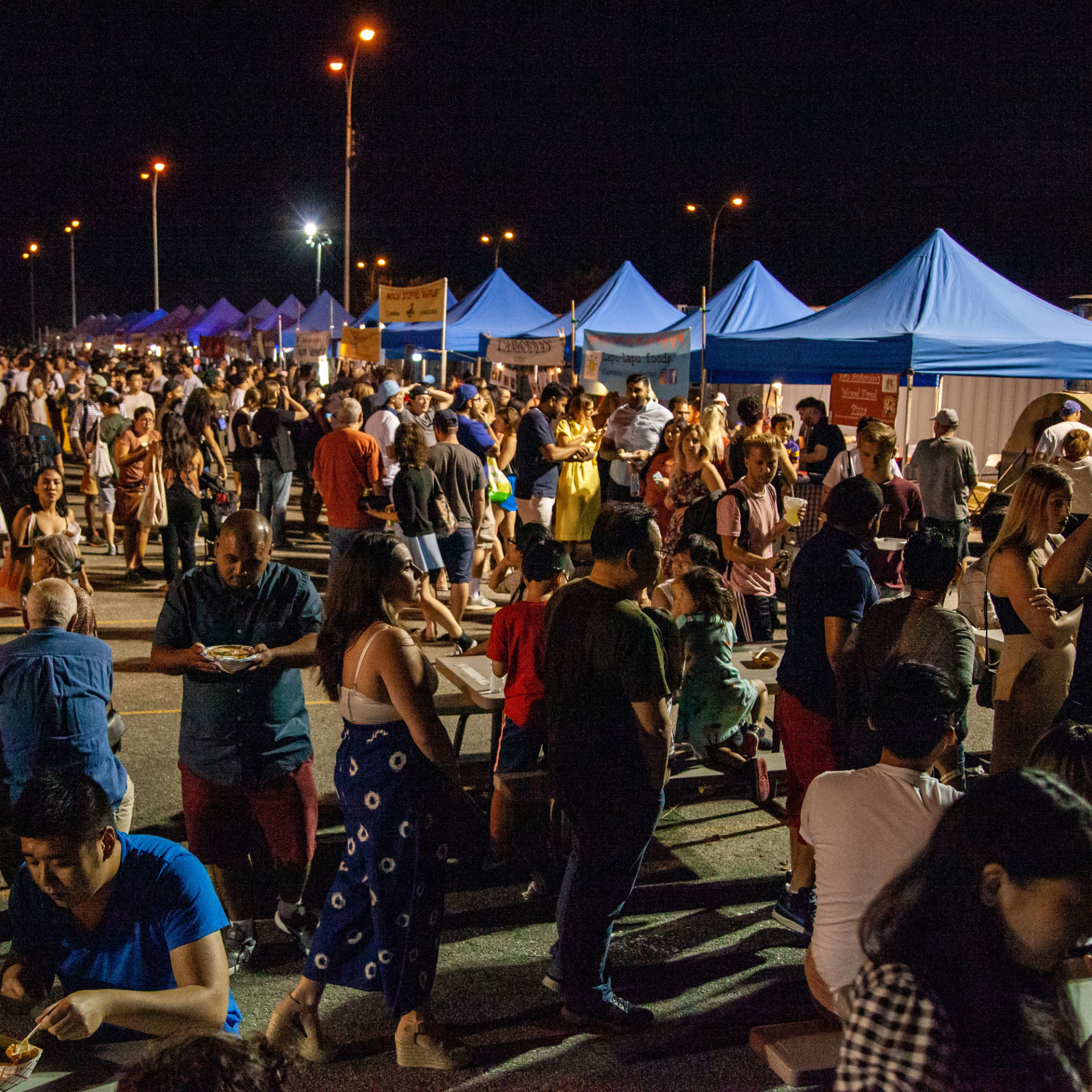 NYC outdoor night market with blue canopy tents, crowds eating and socializing under streetlights, lively cultural food festival featured in daily things to do in NYC any day