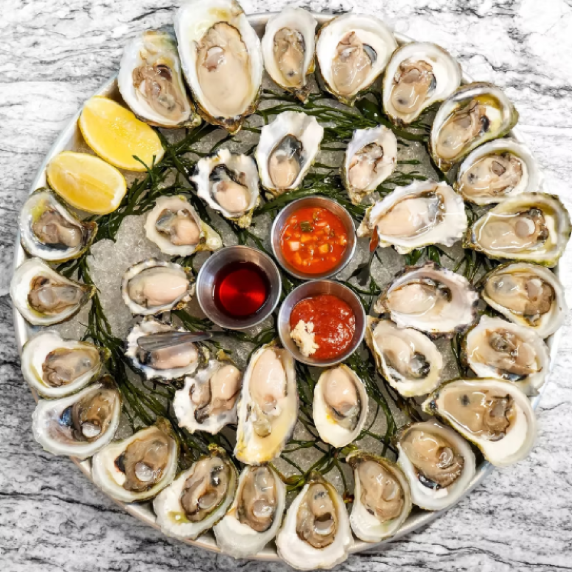 Round platter of freshly shucked NYC oysters on crushed ice with lemon wedges, seaweed garnish, and three sauces in metal cups, seafood dining experience featured in daily things to do in NYC any day