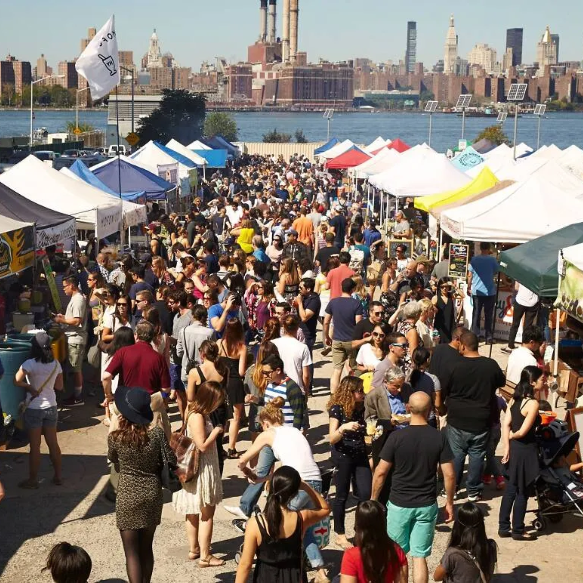 NYC waterfront market with colorful vendor tents and crowds walking by riverfront skyline, lively urban fair featured in daily things to do in NYC any day