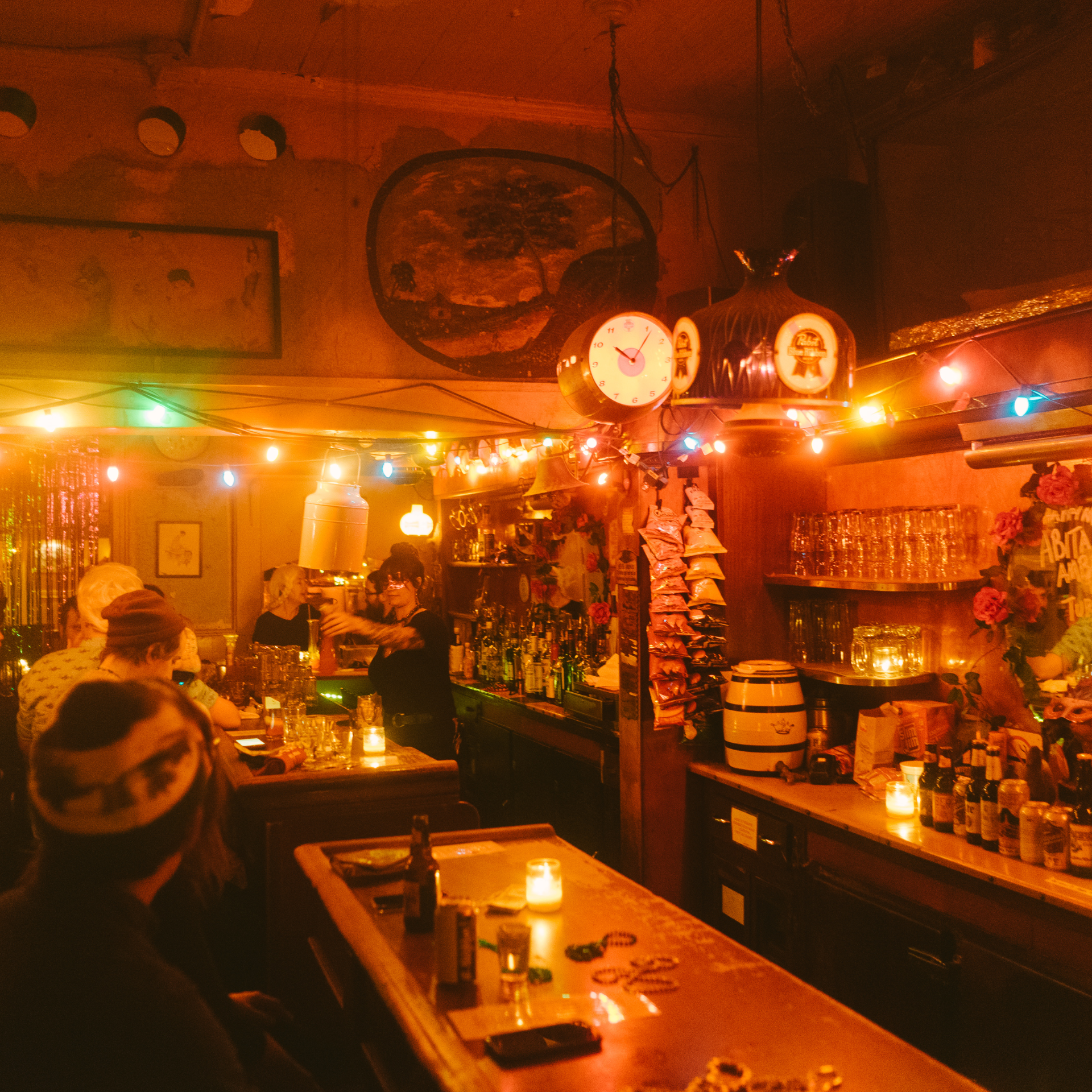 Cozy bar interior with warm lighting, colorful string lights, bartender preparing drinks, patrons seated at counter, bottles and ornaments, clock and wall art décor