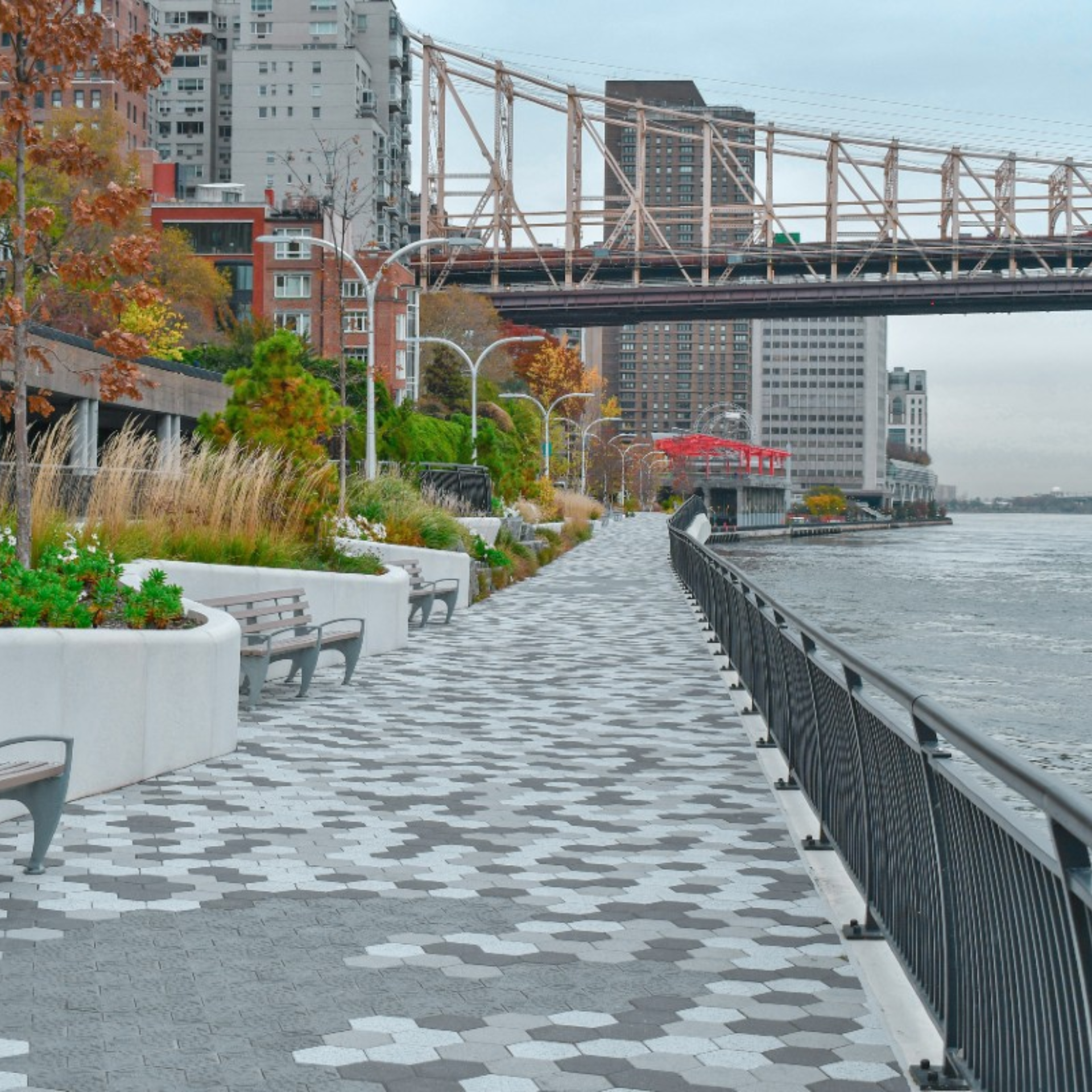 Urban riverside walkway with hexagonal pavement, benches, landscaped planters, modern streetlights, tall buildings, steel bridge, autumn trees along river