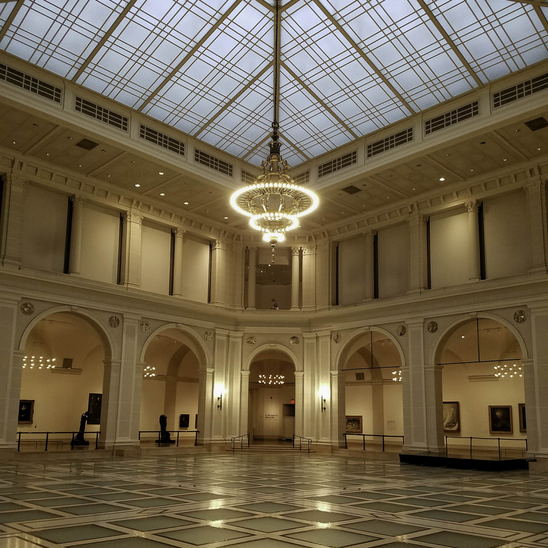 Grand museum hall with vaulted skylight ceiling, tiered chandelier, arches, columns, framed artworks, sculptures, and geometric tile floor