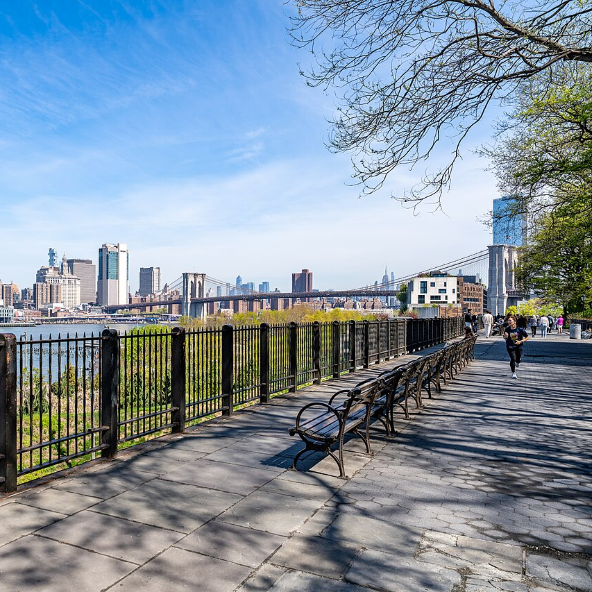 Things to do in NYC any day: Brooklyn Heights Promenade walkway with benches and fence overlooking East River, people walking and jogging, Brooklyn Bridge and Manhattan skyline under clear blue sky