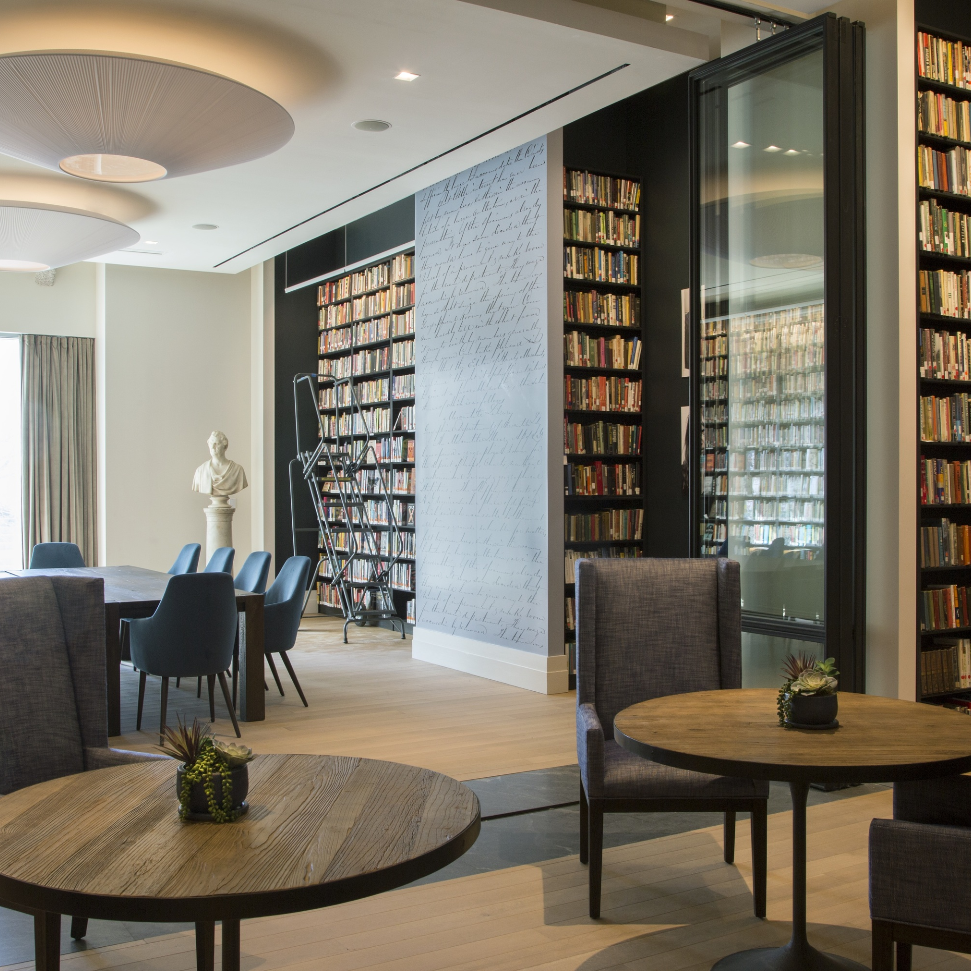 Modern library with tall bookshelves, rolling ladder, bust sculpture, round tables with upholstered chairs, large windows, blue chairs at rectangular table, decorative wall panel
