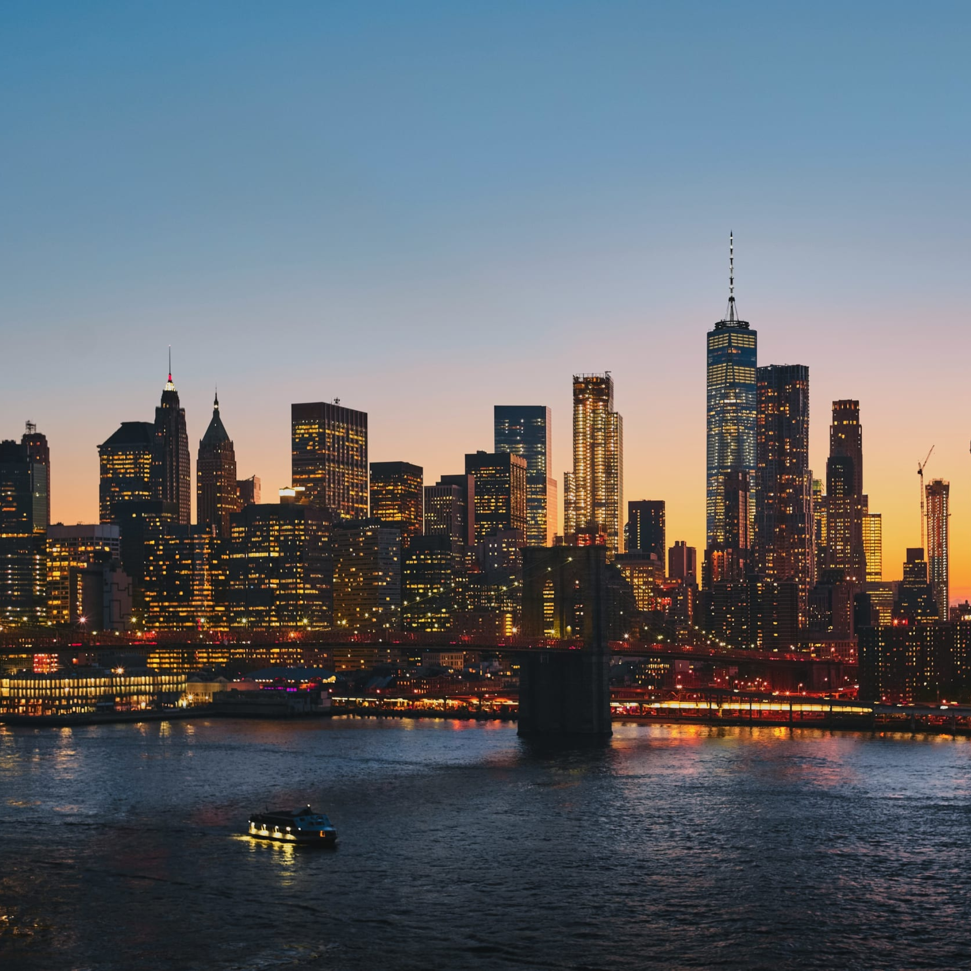City skyline at dusk with illuminated tall buildings, spire tower, bridge over river, sunset sky transitioning orange to deep blue, boat with lights on water