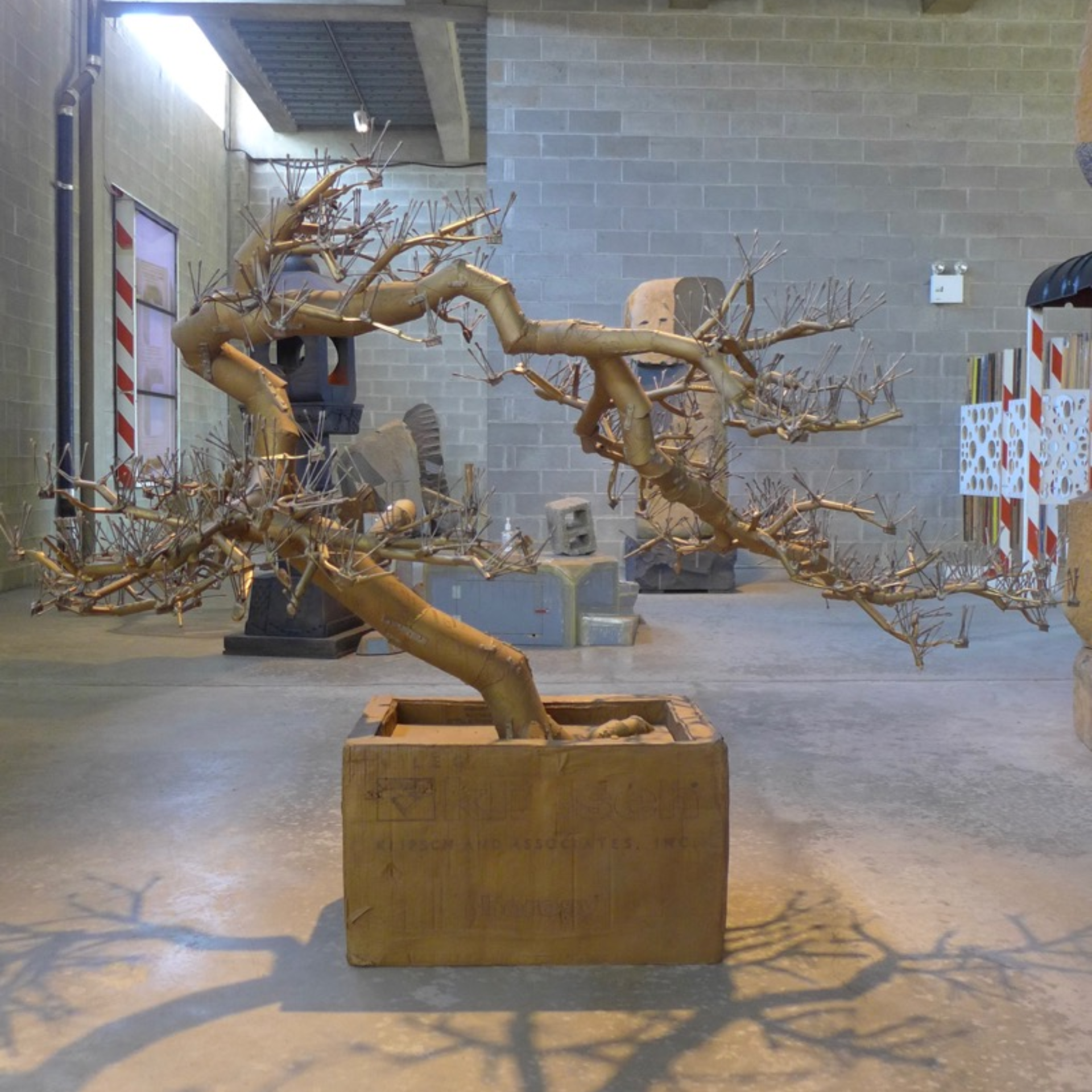 Metal bonsai sculpture with intricate branches in wooden planter, displayed in industrial gallery space with concrete walls and exposed pipes