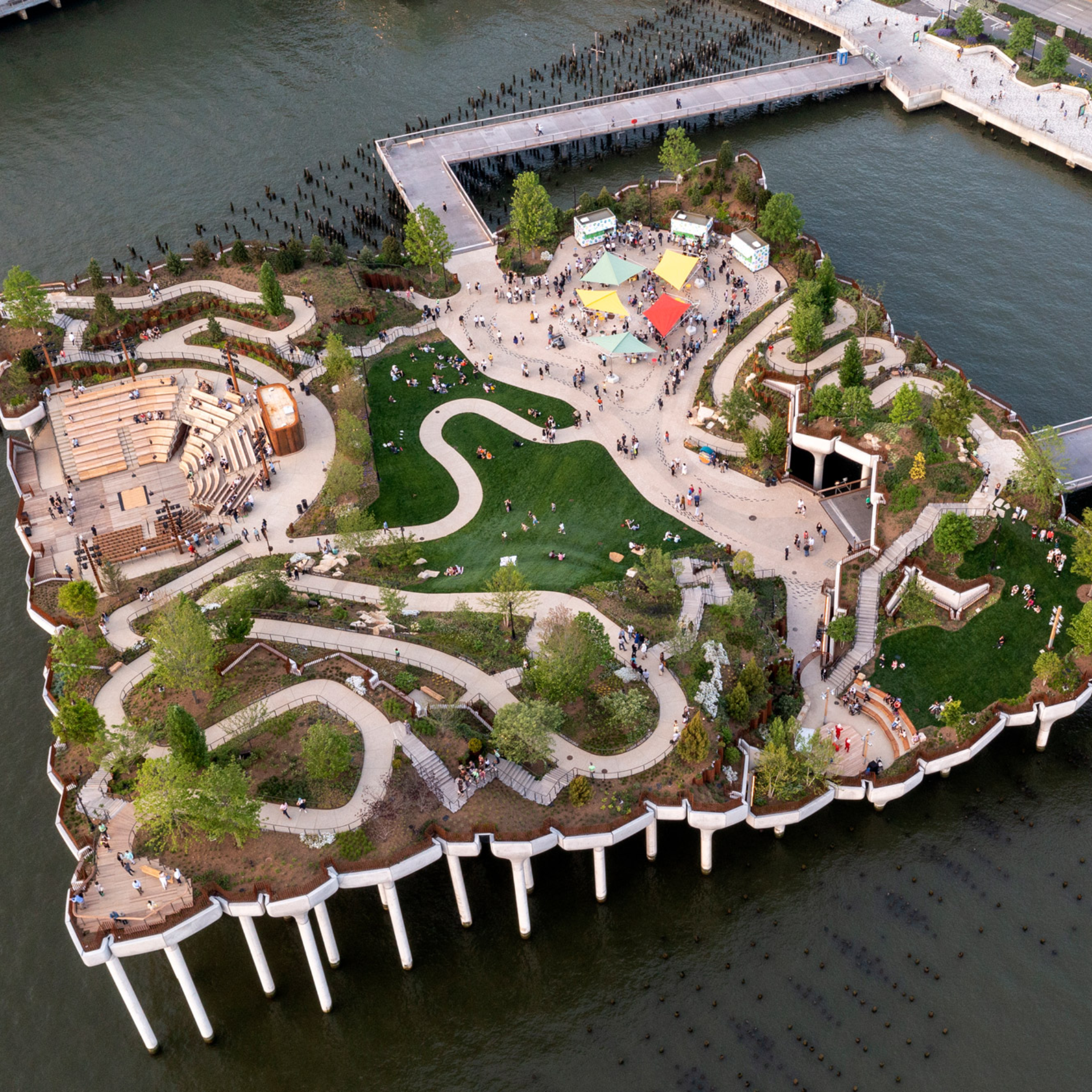 Aerial view of Little Island park on Hudson River with winding pathways, amphitheater seating, canopy structures, landscaped greenery, and city waterfront backdrop