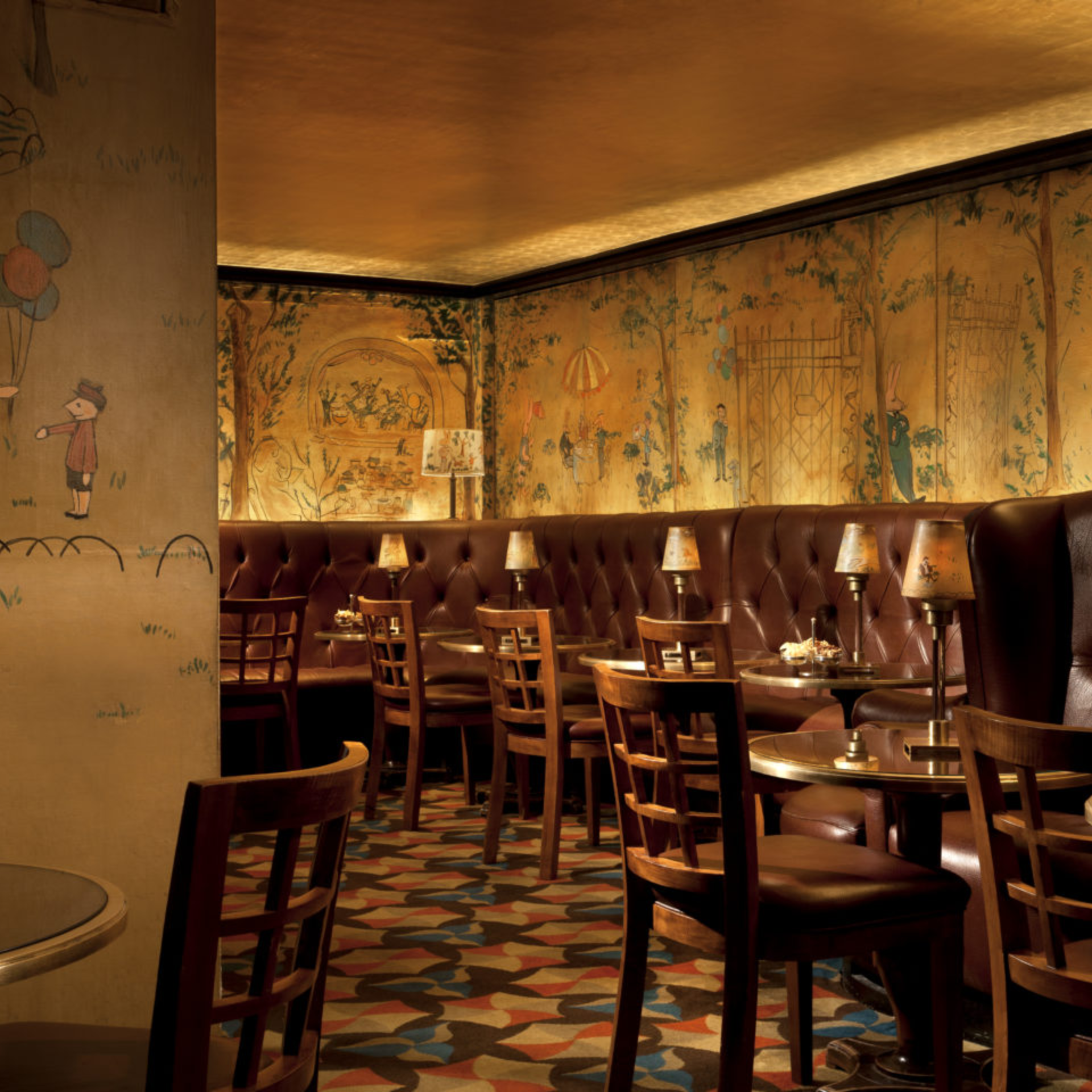 Warmly lit lounge café with brown leather tufted banquette seating, wooden chairs, round tables, lamps with painted shades, whimsical mural walls, colorful geometric carpet