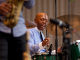 Drummer in striped shirt performing on green drum set with saxophonist in foreground. Things to Do in New York City This August 2026