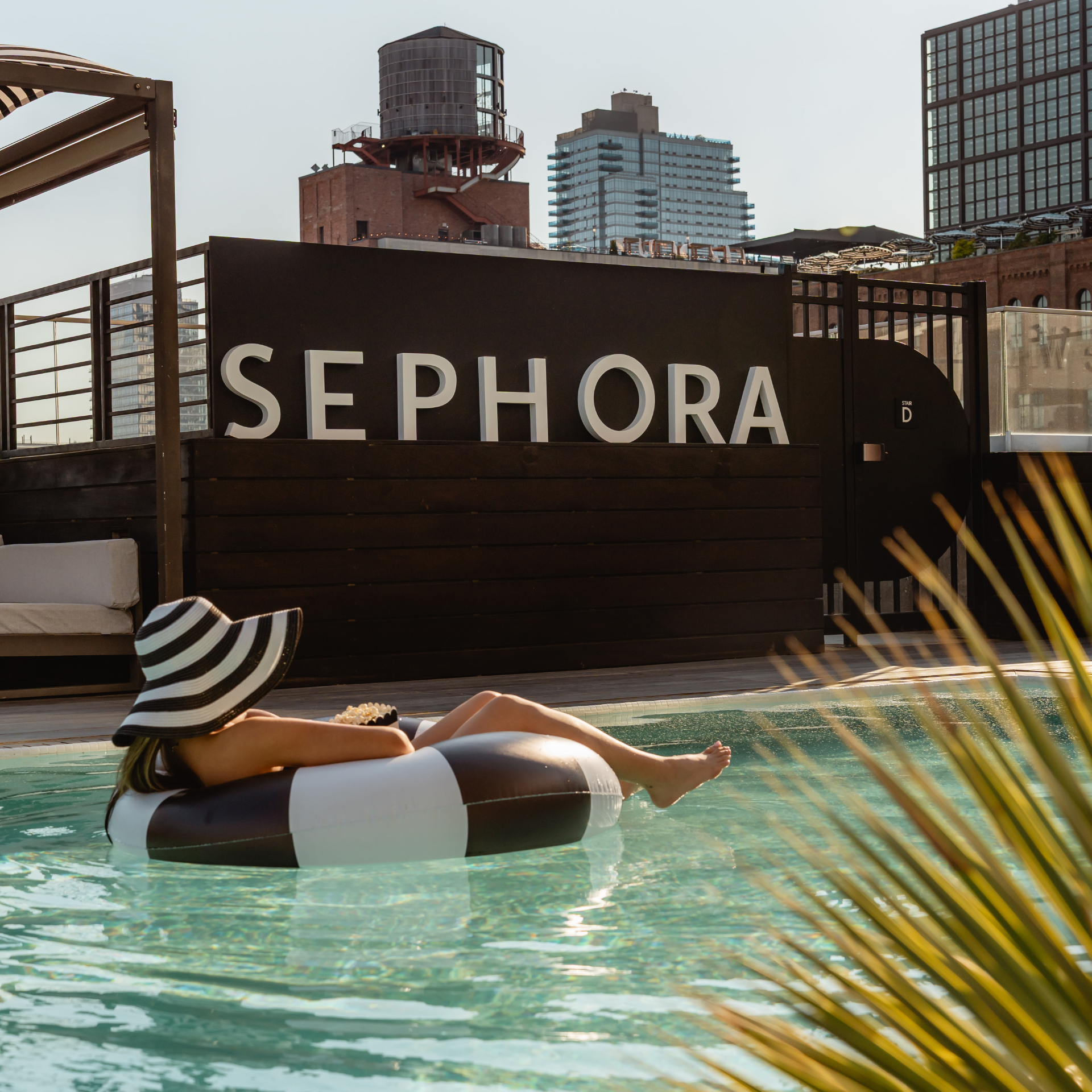 NYC rooftop pool with person relaxing on striped inflatable ring and sun hat, Sephora wall backdrop and city buildings, urban leisure escape featured in daily things to do in NYC any day