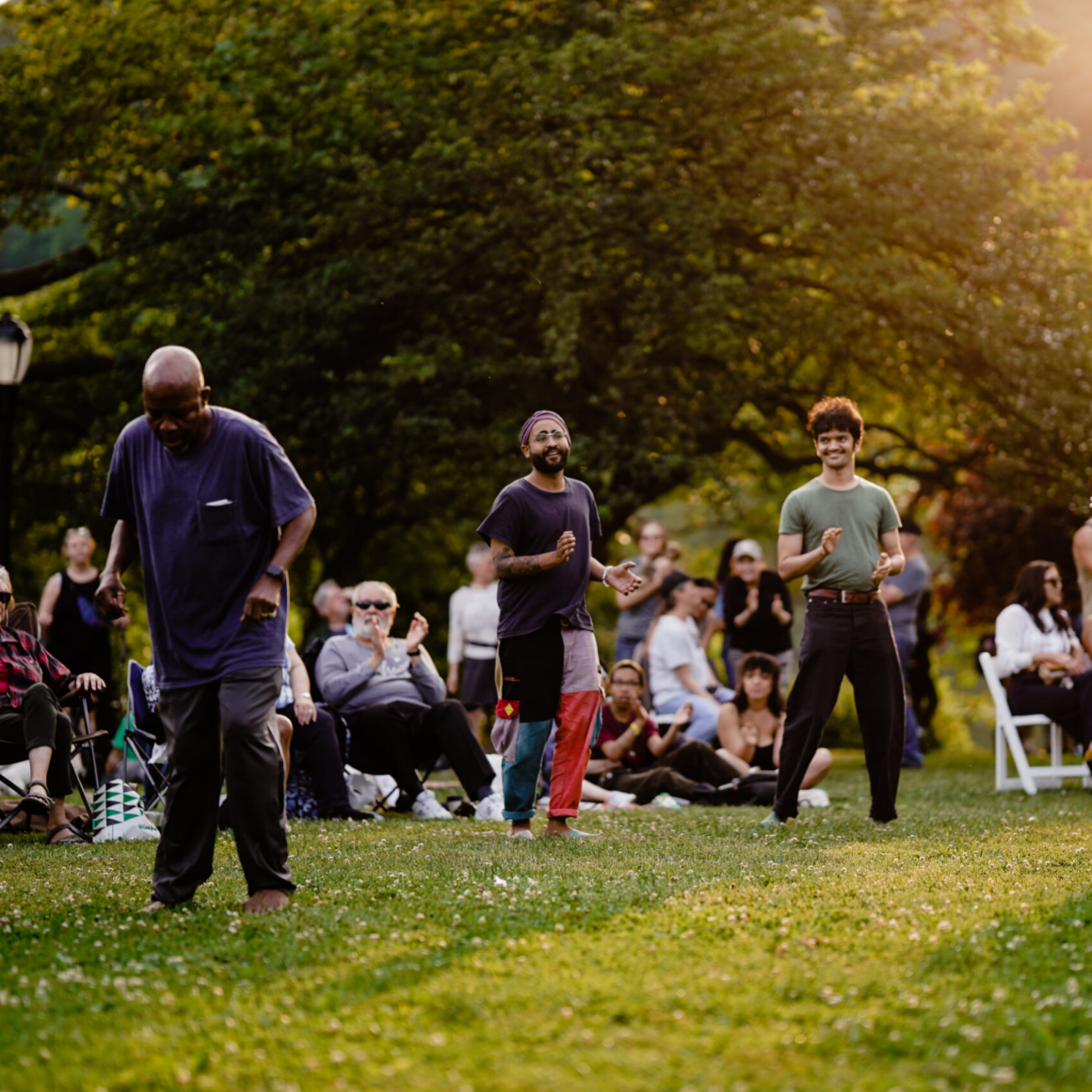 NYC Labor Day park dance performance with three people moving on grass as audience claps under trees and sunlight, outdoor cultural celebration featured in daily things to do in NYC any day