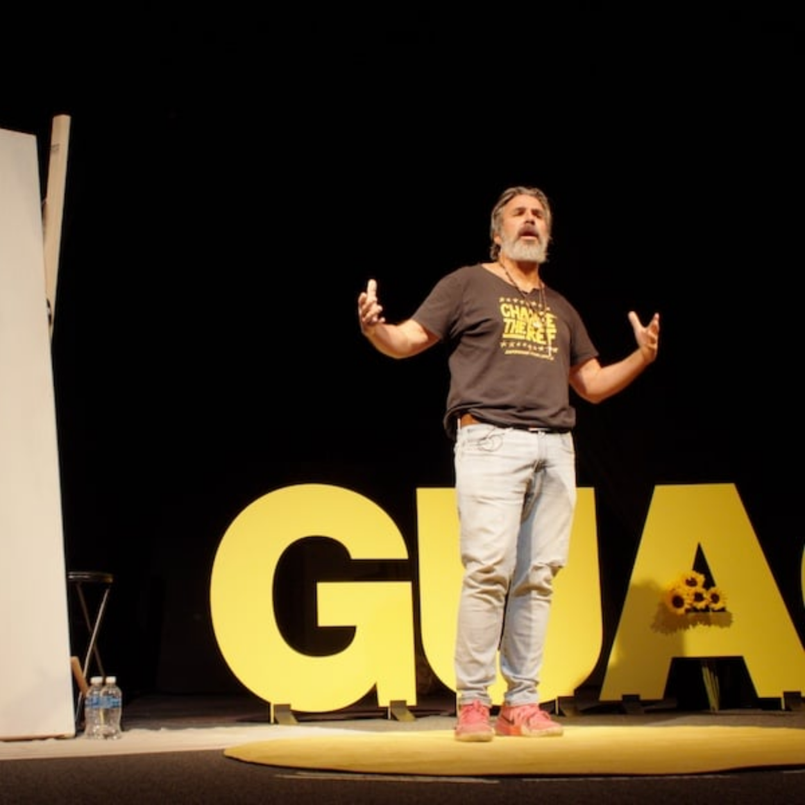 Speaker in black CHANGE THE REF shirt on NYC Labor Day stage with yellow GUAS letters and sunflowers, public talk featured in daily things to do in NYC any day