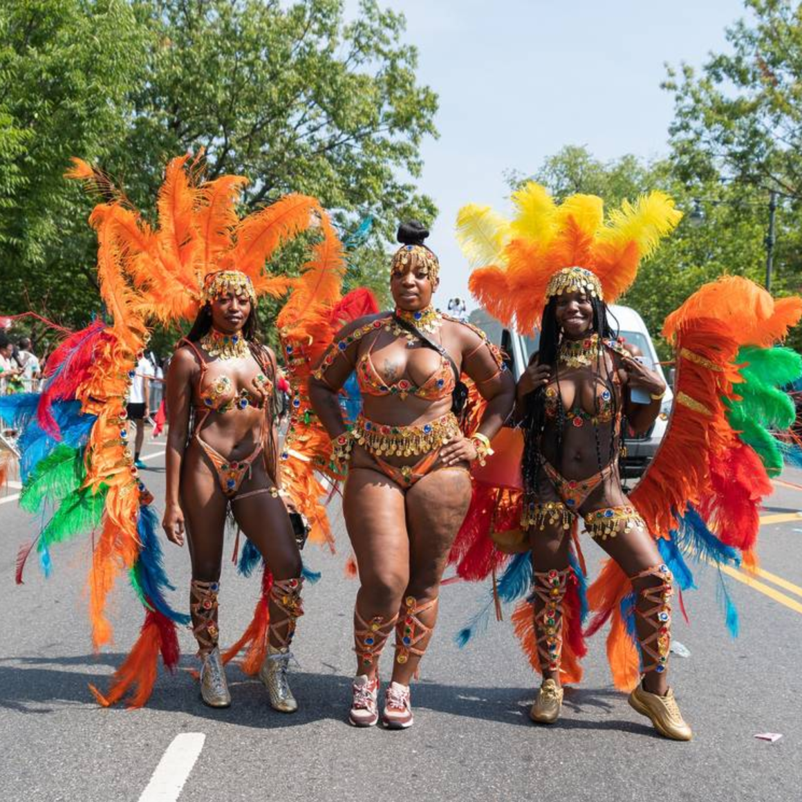 Three NYC parade participants in elaborate feathered costumes with beads, sequins, and gold accents on city street, cultural carnival celebration featured in daily things to do in NYC any day