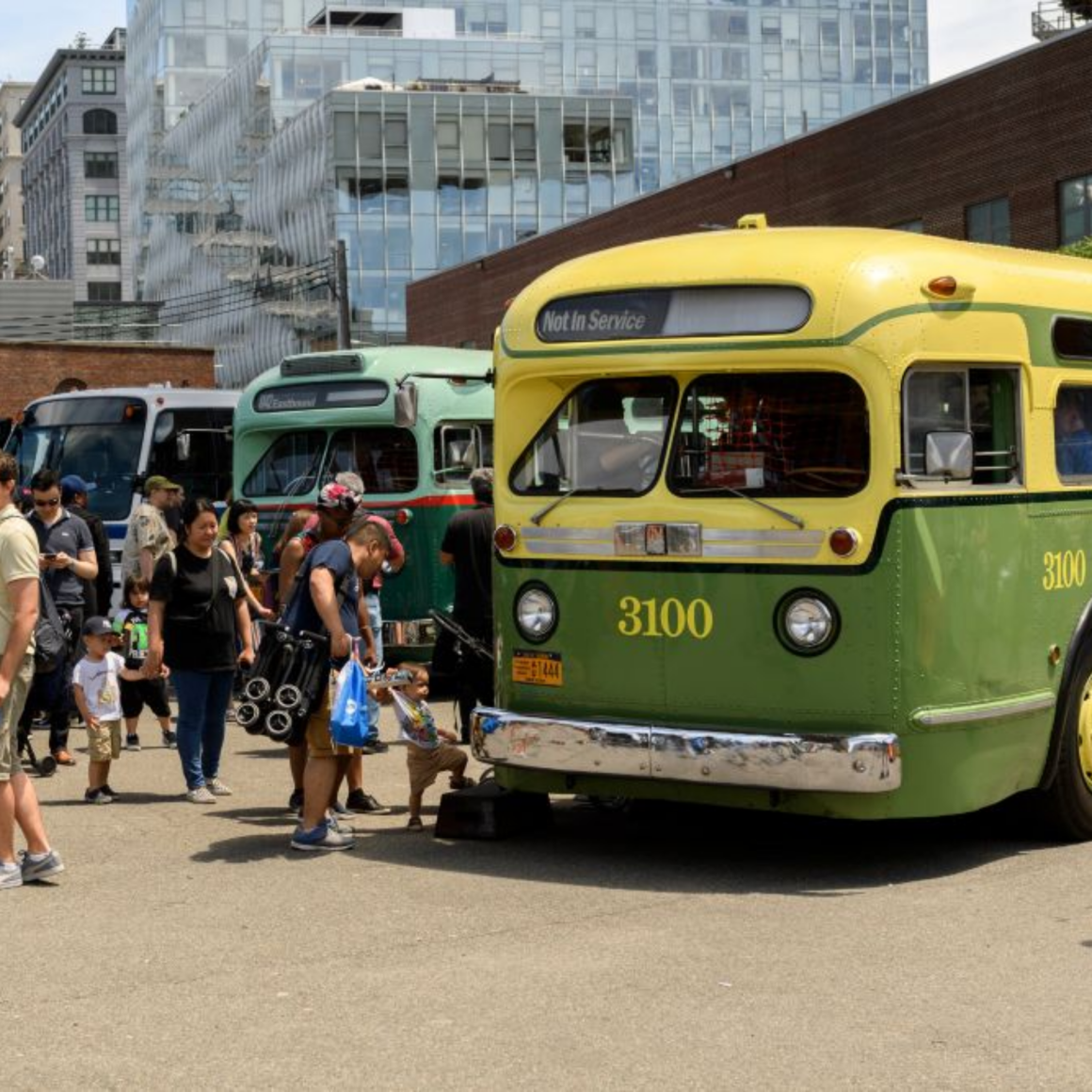 NYC Labor Day vintage bus exhibition with green and yellow bus 3100 and retro fleet against modern glass buildings, transportation history event featured in daily things to do in NYC any day