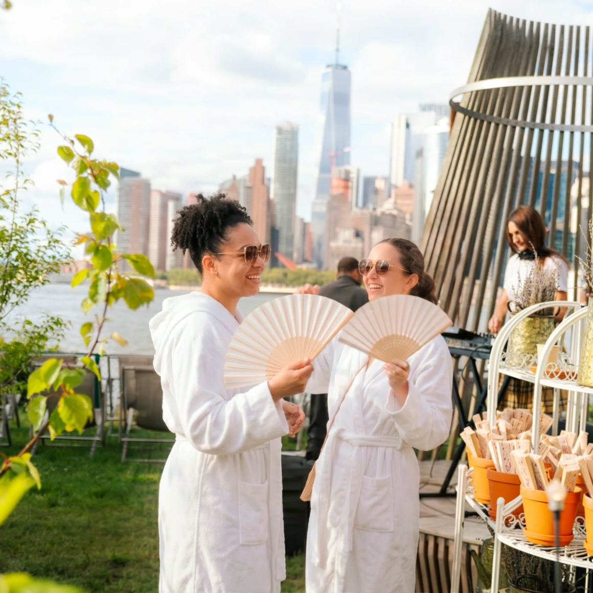 Two people in bathrobes with fans near waterfront skyline