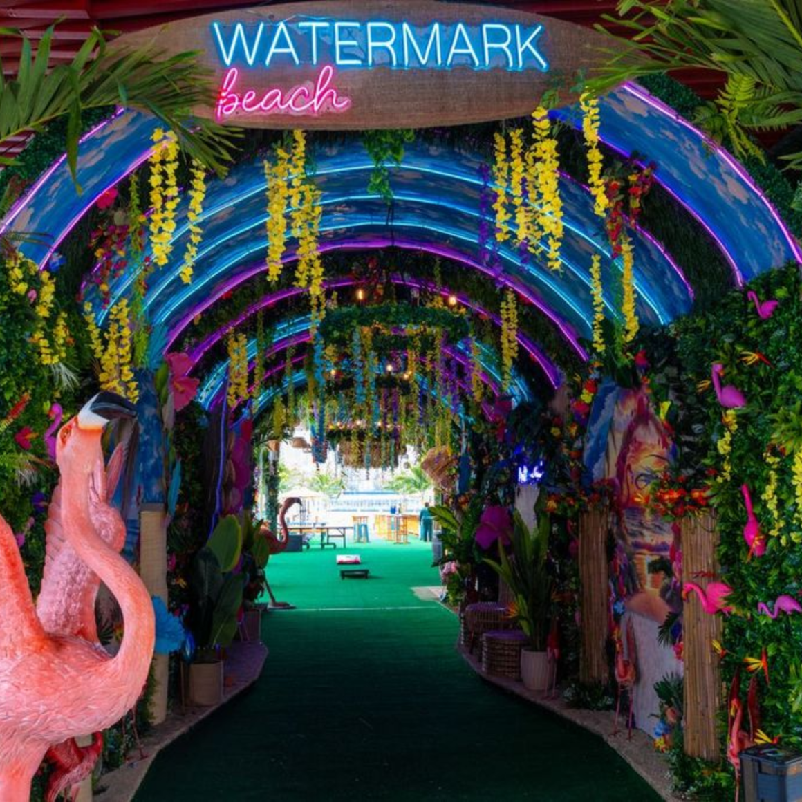 Tropical tunnel with neon “WATERMARK beach” sign, plants, and flamingos