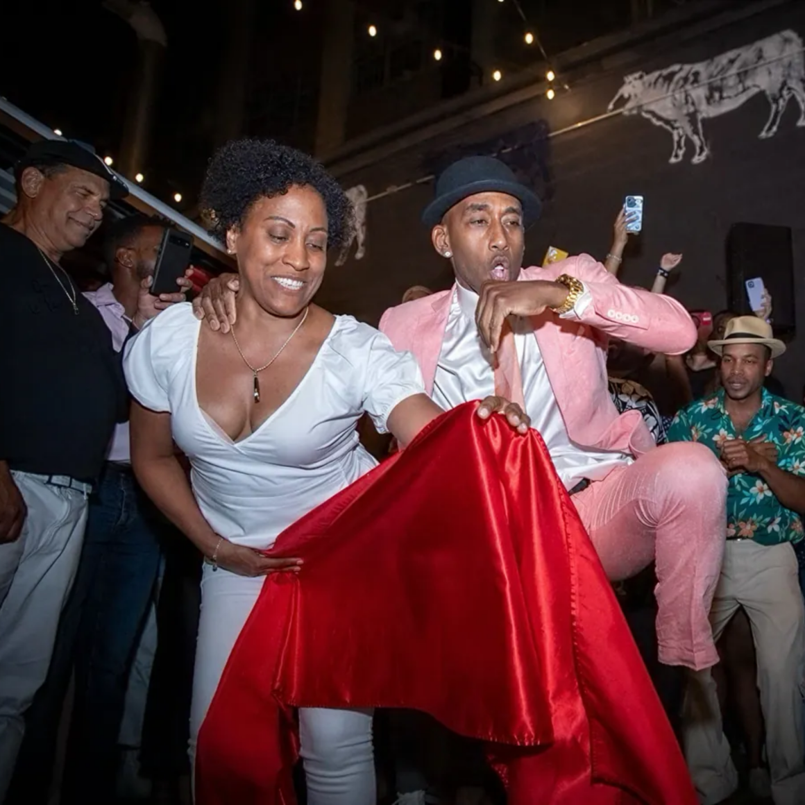 NYC Labor Day street performance with dancer in white holding red cloth and performer in pink suit under string lights, festive cultural celebration featured in daily things to do in NYC any day