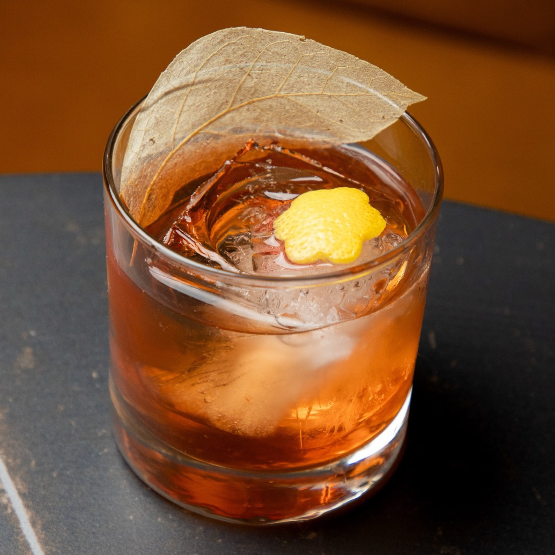 Old‑fashioned glass with amber cocktail, ice cube, and citrus flower garnish