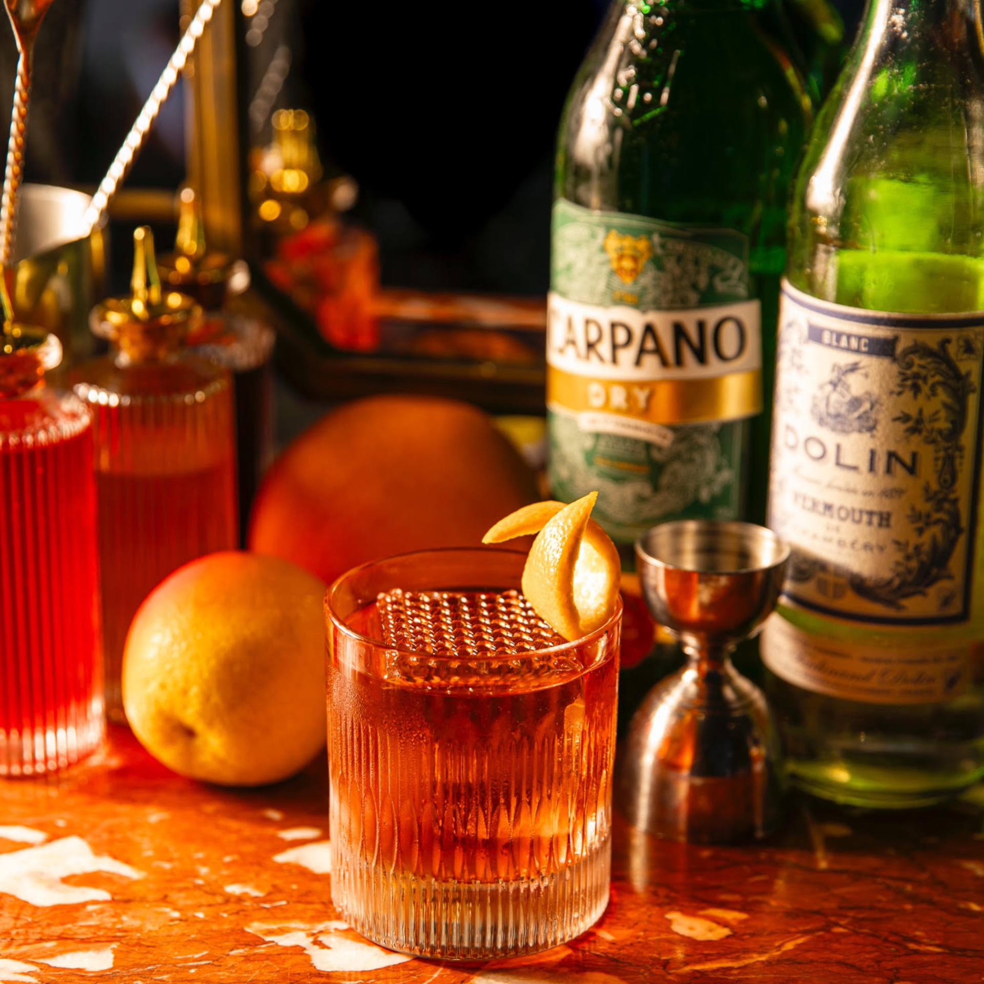 Amber cocktail with lemon twist, vermouth bottles, and bar tools